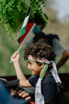 A young child with curly hair raises a flag with red, black, green, and white colors. The child wears a headband and scarf with inscriptions. Sunlight filters through green leaves in the background, creating a natural setting.