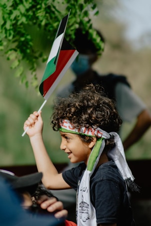 A young child with curly hair raises a flag with red, black, green, and white colors. The child wears a headband and scarf with inscriptions. Sunlight filters through green leaves in the background, creating a natural setting.