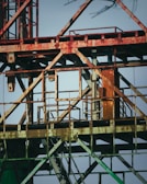 Rusty metallic structure composed of various intersecting beams and frames, possibly part of an industrial site or construction equipment. The metal shows signs of aging and corrosion, with patches of rust and faded paint. The background is a clear blue sky, providing a stark contrast to the aged metal.
