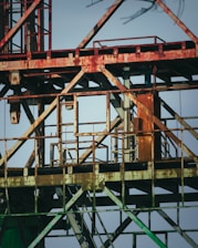 Rusty metallic structure composed of various intersecting beams and frames, possibly part of an industrial site or construction equipment. The metal shows signs of aging and corrosion, with patches of rust and faded paint. The background is a clear blue sky, providing a stark contrast to the aged metal.