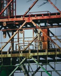 Rusty metallic structure composed of various intersecting beams and frames, possibly part of an industrial site or construction equipment. The metal shows signs of aging and corrosion, with patches of rust and faded paint. The background is a clear blue sky, providing a stark contrast to the aged metal.
