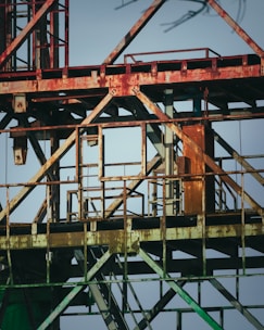Rusty metallic structure composed of various intersecting beams and frames, possibly part of an industrial site or construction equipment. The metal shows signs of aging and corrosion, with patches of rust and faded paint. The background is a clear blue sky, providing a stark contrast to the aged metal.