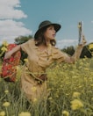 A woman wearing a beige dress and a dark green hat stands in a field of yellow flowers. She holds a red floral-patterned suitcase in one hand and a folded map in the other. The sky is partly cloudy, and there are trees in the background. The scene conveys a sense of adventure or journey.