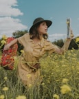 A woman wearing a beige dress and a dark green hat stands in a field of yellow flowers. She holds a red floral-patterned suitcase in one hand and a folded map in the other. The sky is partly cloudy, and there are trees in the background. The scene conveys a sense of adventure or journey.
