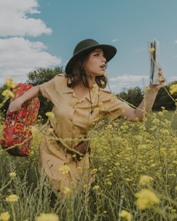 A woman wearing a beige dress and a dark green hat stands in a field of yellow flowers. She holds a red floral-patterned suitcase in one hand and a folded map in the other. The sky is partly cloudy, and there are trees in the background. The scene conveys a sense of adventure or journey.