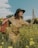 A woman wearing a beige dress and a dark green hat stands in a field of yellow flowers. She holds a red floral-patterned suitcase in one hand and a folded map in the other. The sky is partly cloudy, and there are trees in the background. The scene conveys a sense of adventure or journey.