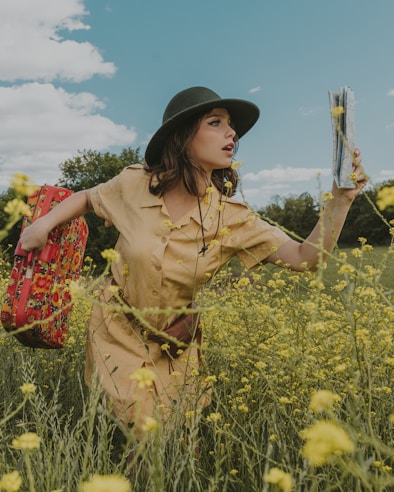 A woman wearing a beige dress and a dark green hat stands in a field of yellow flowers. She holds a red floral-patterned suitcase in one hand and a folded map in the other. The sky is partly cloudy, and there are trees in the background. The scene conveys a sense of adventure or journey.