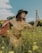 A woman wearing a beige dress and a dark green hat stands in a field of yellow flowers. She holds a red floral-patterned suitcase in one hand and a folded map in the other. The sky is partly cloudy, and there are trees in the background. The scene conveys a sense of adventure or journey.