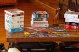 A wooden table is covered with multiple stacks of jigsaw puzzle boxes, each with colorful designs and various piece counts. A completed puzzle depicting a vibrant, urban scene with neon lights and a rainy street is displayed on the table. A small wooden giraffe statue is positioned next to one of the stacks. In the lower left corner, part of a dog’s face is visible looking up at the table.