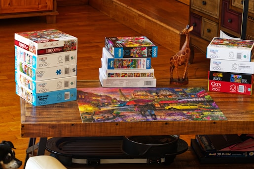 A wooden table is covered with multiple stacks of jigsaw puzzle boxes, each with colorful designs and various piece counts. A completed puzzle depicting a vibrant, urban scene with neon lights and a rainy street is displayed on the table. A small wooden giraffe statue is positioned next to one of the stacks. In the lower left corner, part of a dog&rsquo;s face is visible looking up at the table.
