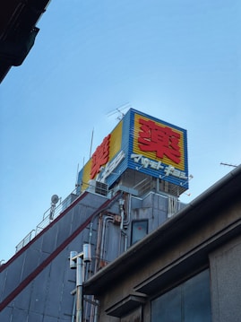 A building rooftop with a colorful outdoor advertisement displaying large, bold red and yellow characters against a blue background. Various antennas and metal pipes are visible, along with the grey, industrial exterior of the building.