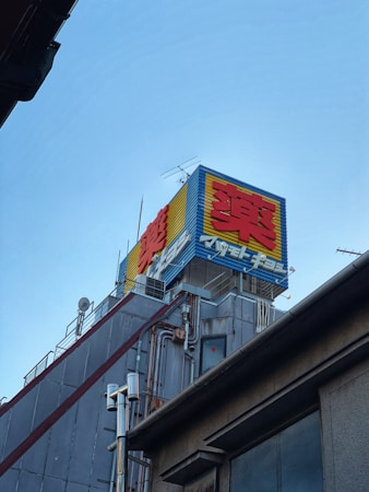A building rooftop with a colorful outdoor advertisement displaying large, bold red and yellow characters against a blue background. Various antennas and metal pipes are visible, along with the grey, industrial exterior of the building.