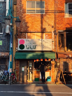 A small eatery with a brick facade is bathed in warm sunlight. The sign above the entrance has Japanese characters, and a man is standing at the counter. Bright paper lanterns are hanging above the entrance, and bicycles are parked alongside the building.