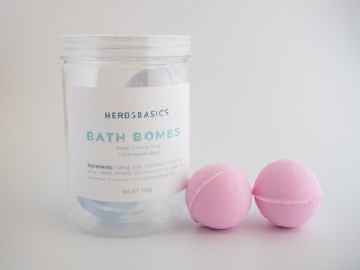 A clear container labeled 'HERBS BASICS BATH BOMBS' stands beside two pink bath bombs. The label indicates the product is handmade in Hong Kong and lists ingredients including baking soda, citric acid, and sweet almond oil. The background is a simple, light gray.