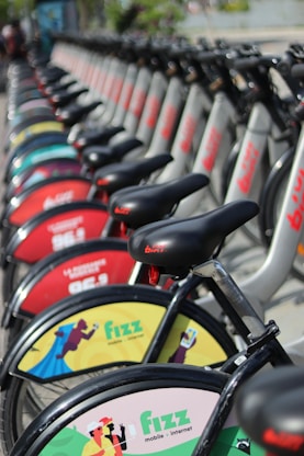 A row of bicycles parked in a line, each featuring advertisements on their wheel covers. The bicycles are predominantly silver with black seats and handlebars, and the advertisements are colorful, including logos and creative illustrations. The focus of the image is on the front wheels with branded covers, and the background fades with more bikes continuing in perspective.