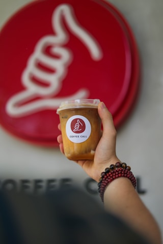 A hand holding a cold coffee drink in a plastic cup with a logo featuring a hand gesture in front of a matching larger logo on a wall. The person is wearing a beaded bracelet.