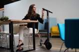 A woman seated at a wooden table in a modern indoor setting with a white wall background. She wears a black top, white pants, and black shoes, resting her arm on a black electric scooter marked with the brand 'MEARTH'. The setting is accented with colorful chairs, including a blue and a mustard yellow chair, alongside a small green plant on the table.
