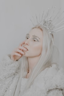 A person with long, pale blond hair is wearing a textured light gray coat and an ornate silver crown with pointed elements. The person's eyebrows are adorned with silver or crystal embellishments. The overall aesthetic is ethereal and wintry.