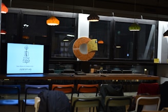 A modern indoor space with a series of pendant lights hanging from the ceiling, casting a warm glow. In front of a row of large windows, there's a table with a monitor showing a simplistic drawing and text. Nearby, several colorful chairs are arranged, and a circular object adorned with handwritten notes is on display.