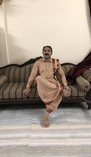 A man with a mustache sits cross-legged on a striped sofa. He is wearing traditional attire consisting of a beige kurta and dhoti with a brown sash. The setting appears to be indoors with a plain wall and some draped fabric enhancing the furniture.