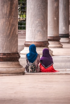 Two individuals wearing colorful clothing and head coverings sit together between large stone columns. The setting appears to be a historic or architectural site with a serene and contemplative atmosphere.
