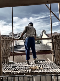 A construction worker stands on a bamboo scaffold, looking towards the city skyline. The scene includes rooftops, buildings, and a partly cloudy sky. The worker is dressed in blue pants, a helmet, and gloves, with tools around his feet.