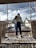 A construction worker stands on a bamboo scaffold, looking towards the city skyline. The scene includes rooftops, buildings, and a partly cloudy sky. The worker is dressed in blue pants, a helmet, and gloves, with tools around his feet.