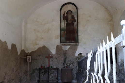 A rustic interior scene with a religious motif, featuring a wall-mounted painting of a bearded figure holding a staff. Below the painting is a simple altar with a wooden cross, framed picture, candles, and small vase. The setting is enclosed by aged, textured walls and a metal fence.
