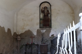 A rustic interior scene with a religious motif, featuring a wall-mounted painting of a bearded figure holding a staff. Below the painting is a simple altar with a wooden cross, framed picture, candles, and small vase. The setting is enclosed by aged, textured walls and a metal fence.