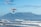 A powered hang glider with an orange wing is flying over a coastline with waves gently crashing onto the shore. In the background, a prominent flat-topped mountain rises under a bright blue sky with light clouds.