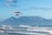 A powered hang glider with an orange wing is flying over a coastline with waves gently crashing onto the shore. In the background, a prominent flat-topped mountain rises under a bright blue sky with light clouds.