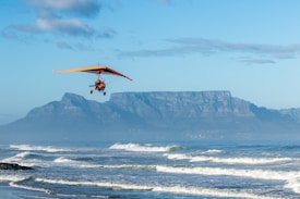 A powered hang glider with an orange wing is flying over a coastline with waves gently crashing onto the shore. In the background, a prominent flat-topped mountain rises under a bright blue sky with light clouds.