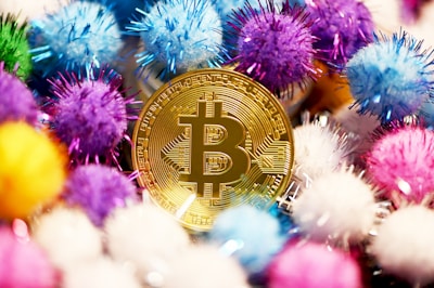 A gold-colored Bitcoin coin is surrounded by colorful decorative pom-poms in various shades of blue, purple, pink, yellow, and white. The arrangement creates a vibrant and playful atmosphere.