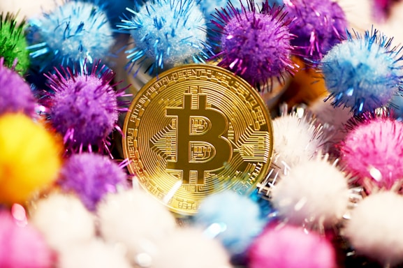 A gold-colored Bitcoin coin is surrounded by colorful decorative pom-poms in various shades of blue, purple, pink, yellow, and white. The arrangement creates a vibrant and playful atmosphere.