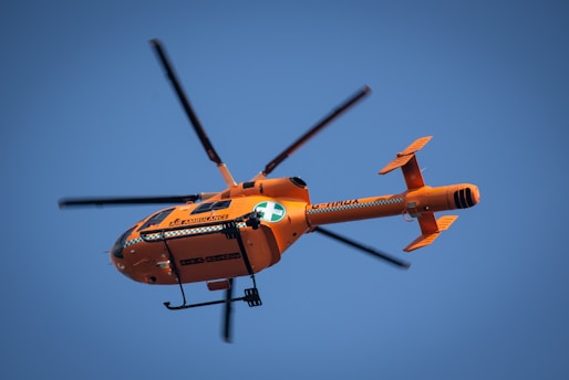 An orange air ambulance helicopter is flying in a clear blue sky. The helicopter has a green and white cross emblem, typical of medical transport. Its rotors are in motion, and there are visible markings and text on the side.