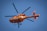 An orange air ambulance helicopter is flying in a clear blue sky. The helicopter has a green and white cross emblem, typical of medical transport. Its rotors are in motion, and there are visible markings and text on the side.