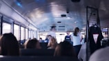 Inside a bus, young people are interacting with each other. Several passengers are seated while a few are standing and talking. On the right, some clothing items, including a t-shirt, hang from a rack.