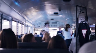 Inside a bus, young people are interacting with each other. Several passengers are seated while a few are standing and talking. On the right, some clothing items, including a t-shirt, hang from a rack.