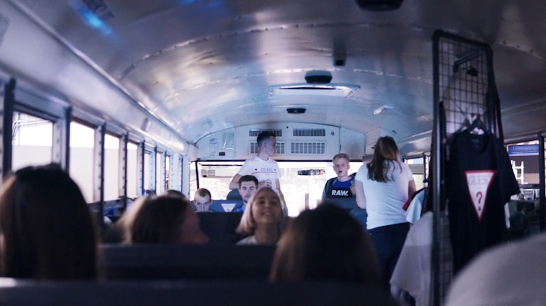 Inside a bus, young people are interacting with each other. Several passengers are seated while a few are standing and talking. On the right, some clothing items, including a t-shirt, hang from a rack.