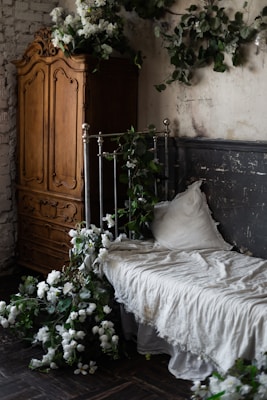 A rustic bedroom scene features a vintage metal bed frame adorned with lush greenery and white flowers. The bedding looks soft and slightly wrinkled, giving a cozy, lived-in feel. Against the weathered, textured walls is a large wooden armoire with intricate carvings, adding a touch of elegance to the room.