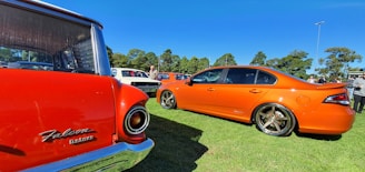Several vintage and modern cars are parked on a grassy area under a clear blue sky. An orange sedan with custom wheels is prominently displayed alongside a classic red Falcon car. A few people are mingling around, possibly at a car show or exhibition.