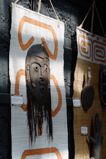 A wooden tribal mask with intricate carvings is mounted on a woven mat with orange patterns. The mask includes long hair-like material hanging down. Next to it, another mat displays a carved wooden object. Both items appear to be cultural or traditional artifacts displayed in an exhibit setting.