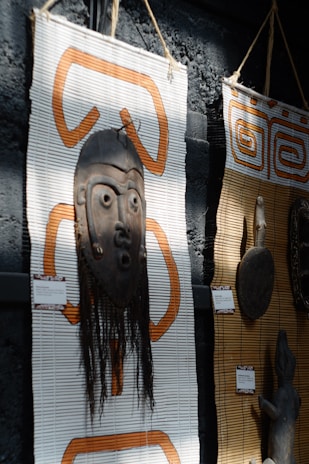 A wooden tribal mask with intricate carvings is mounted on a woven mat with orange patterns. The mask includes long hair-like material hanging down. Next to it, another mat displays a carved wooden object. Both items appear to be cultural or traditional artifacts displayed in an exhibit setting.