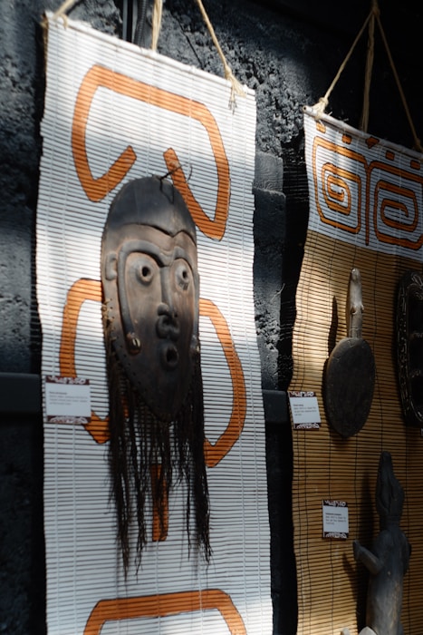 A wooden tribal mask with intricate carvings is mounted on a woven mat with orange patterns. The mask includes long hair-like material hanging down. Next to it, another mat displays a carved wooden object. Both items appear to be cultural or traditional artifacts displayed in an exhibit setting.