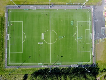An aerial view of a green soccer field with white boundary lines, perimeter fencing, and multiple players scattered across the pitch. Adjacent areas include pathways and some surrounding greenery.