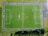 An aerial view of a green soccer field with white boundary lines, perimeter fencing, and multiple players scattered across the pitch. Adjacent areas include pathways and some surrounding greenery.