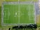 An aerial view of a green soccer field with white boundary lines, perimeter fencing, and multiple players scattered across the pitch. Adjacent areas include pathways and some surrounding greenery.
