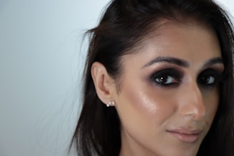 Close-up portrait of a person with dark eye makeup and light nude lipstick. Their hair is dark and styled simply, with a focus on the eyes. The background is a plain, pale color, emphasizing the subject's face.
