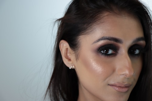Close-up portrait of a person with dark eye makeup and light nude lipstick. Their hair is dark and styled simply, with a focus on the eyes. The background is a plain, pale color, emphasizing the subject's face.