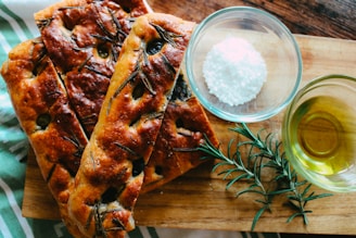 Freshly baked focaccia bread topped with rosemary is arranged on a wooden board. Nearby, a small glass bowl holds coarse salt, and another bowl contains olive oil. A sprig of rosemary is also placed next to the bowls.